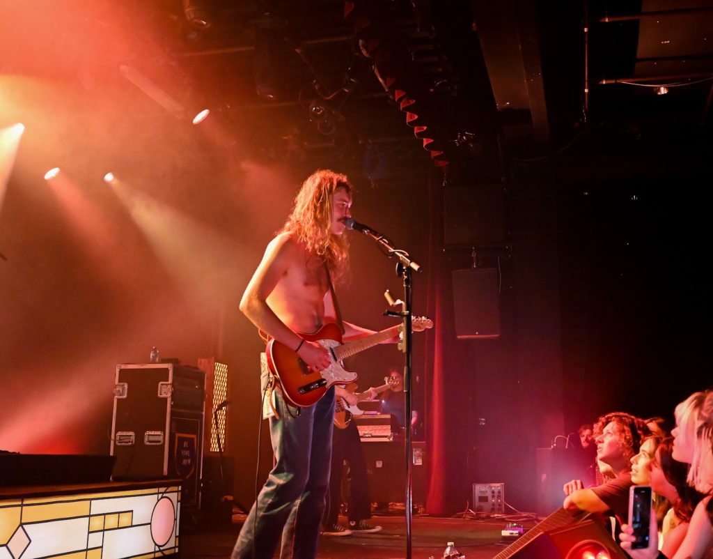 A shirtless man with long hair plays guitar and sings on stage in front of a crowd.