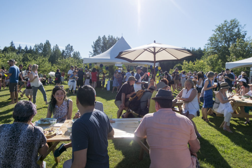 An outdoor food event on a farm with several people standing or sitting at picnic tables eating and drinking. 