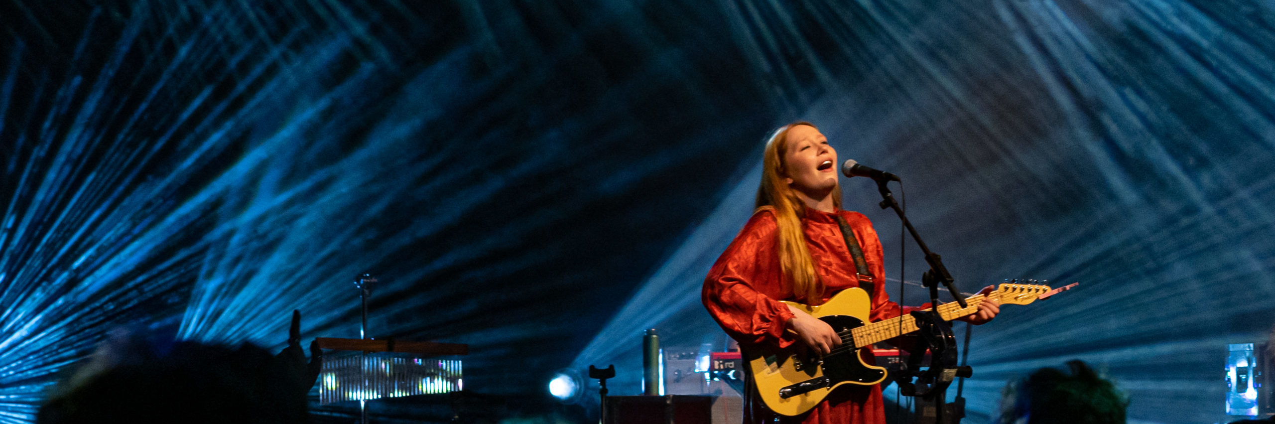 Audience view of a female singer dressed in a red dress playing guitar on stage lit by myltipple reflectors casting blue rays towards the audience