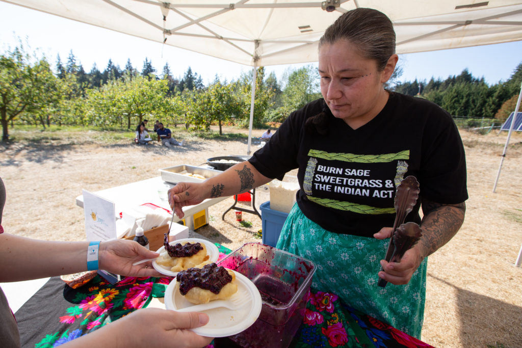A woman places a spoonful of jam on bannock at an outdoor culinary event. Her t-shirt says, "Burn sage sweetgrass & the Indian Act". 