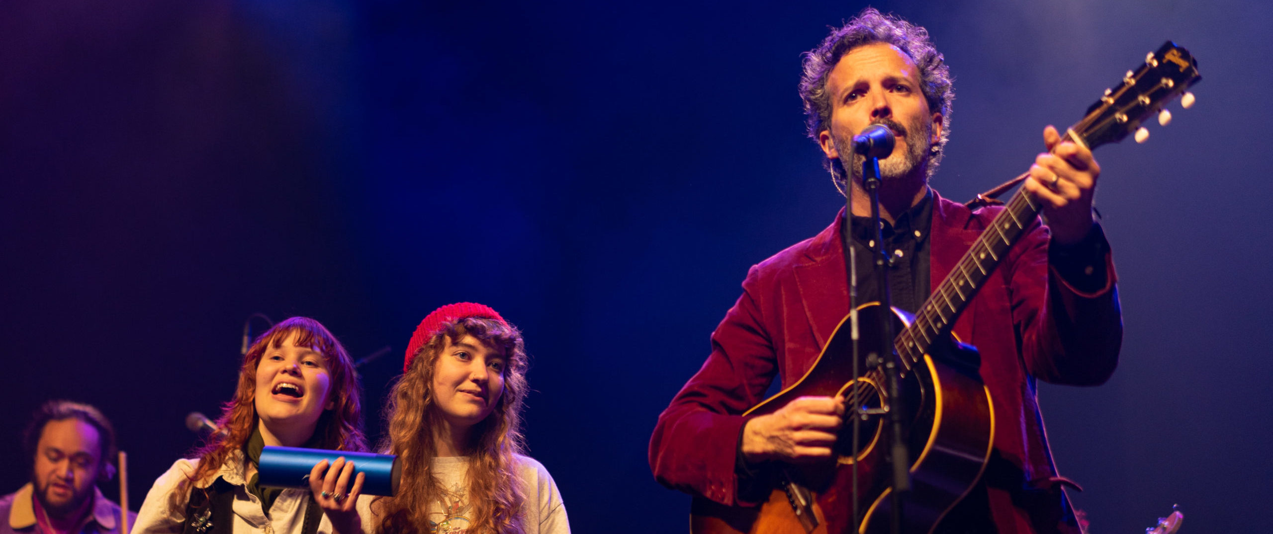 A singer playing guitar and singing with two percussion musicians on his left side on a blue lit stage