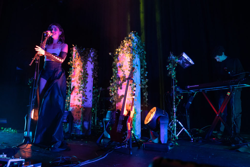 Singer Kimbra is on the left side of the photo singing on a stage. Behind her and to the right is a man playing a synthesizer.