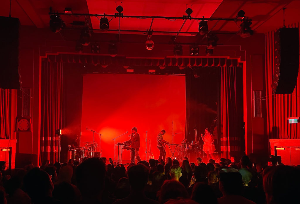 Montreal-based Chiiild, performs in a theatre, on stage in front of a crowd. The theatre is lit up with red lights.