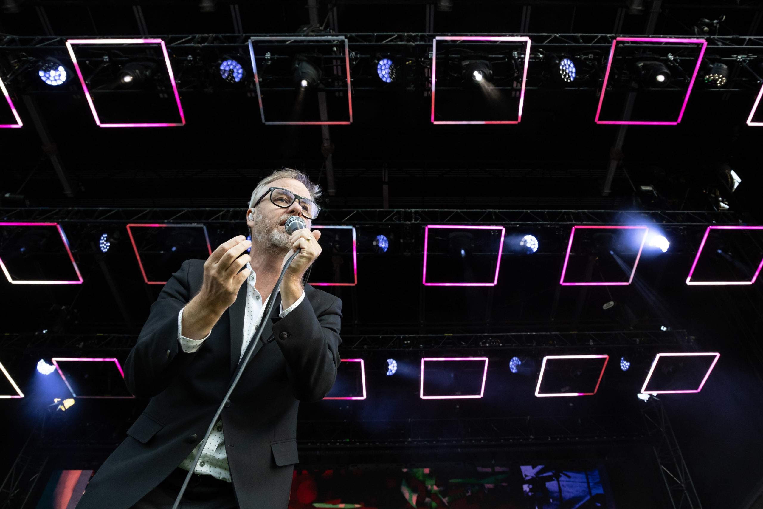 The front man from The National, Matt Berninger, stands under square lights on the stage at Deer Lake Park