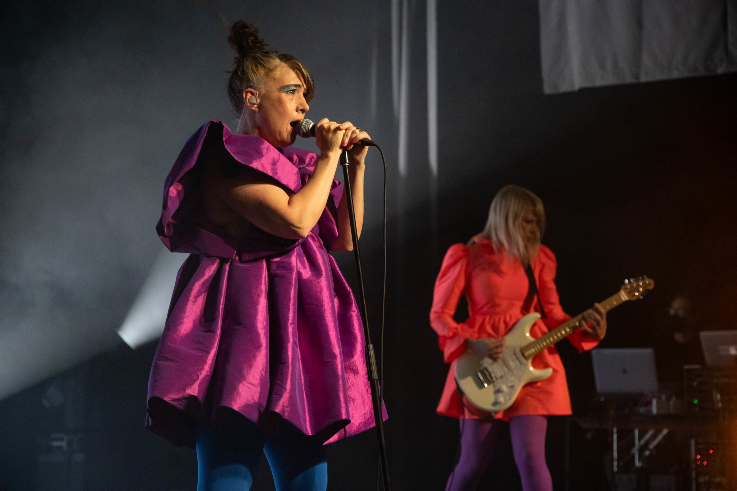 The front woman for Le Tigre is on stage at the Commodore in a shiny purple dress, with another member of the band in an orange dress behind her