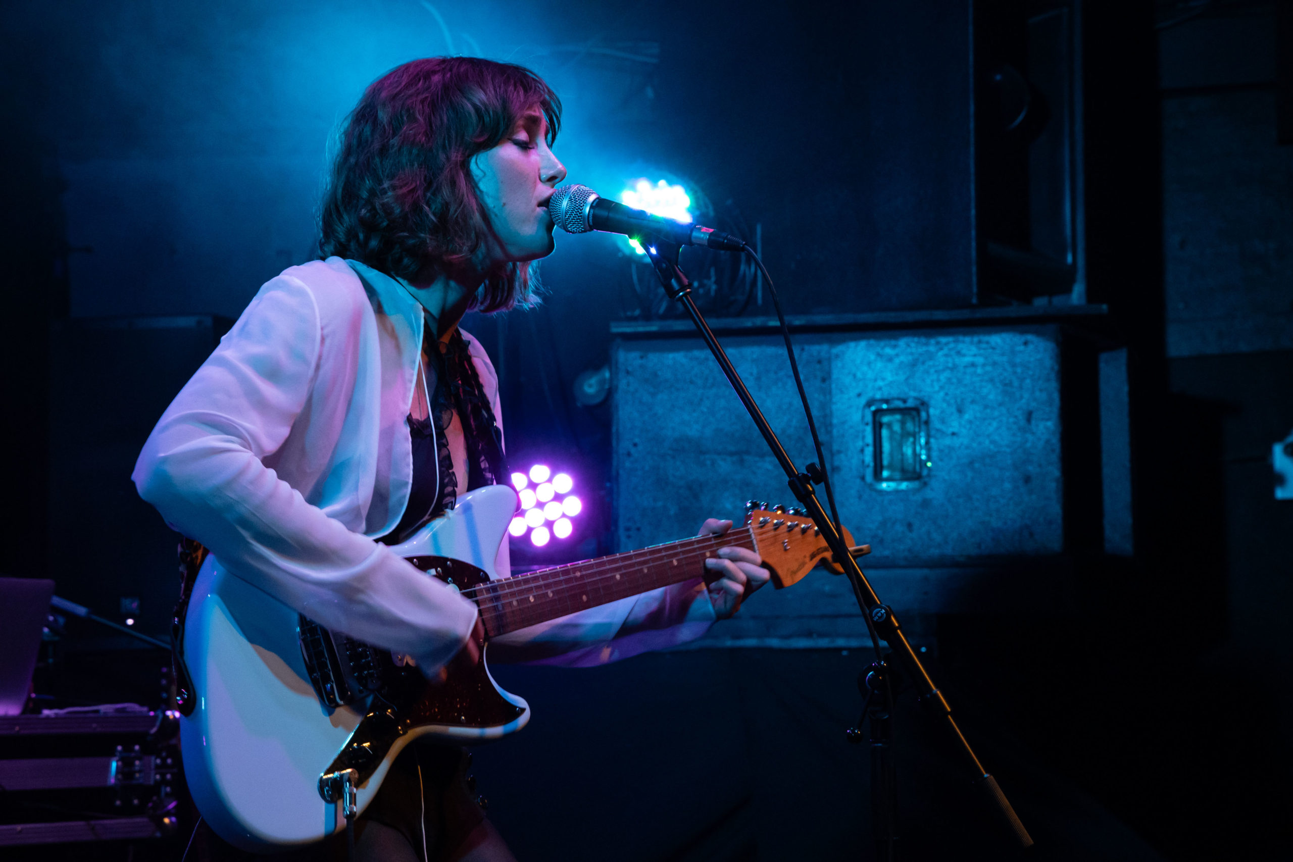 Zella Day sings into the microphone while playing the guitar bathed in blue light