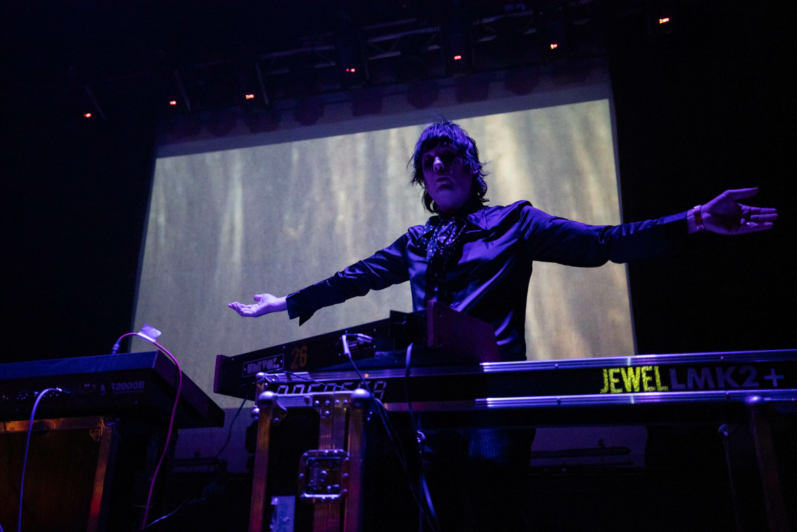 Johnny Jewel lifts his arms as he stands in front of a screen on the stage of the Rickshaw Theatre