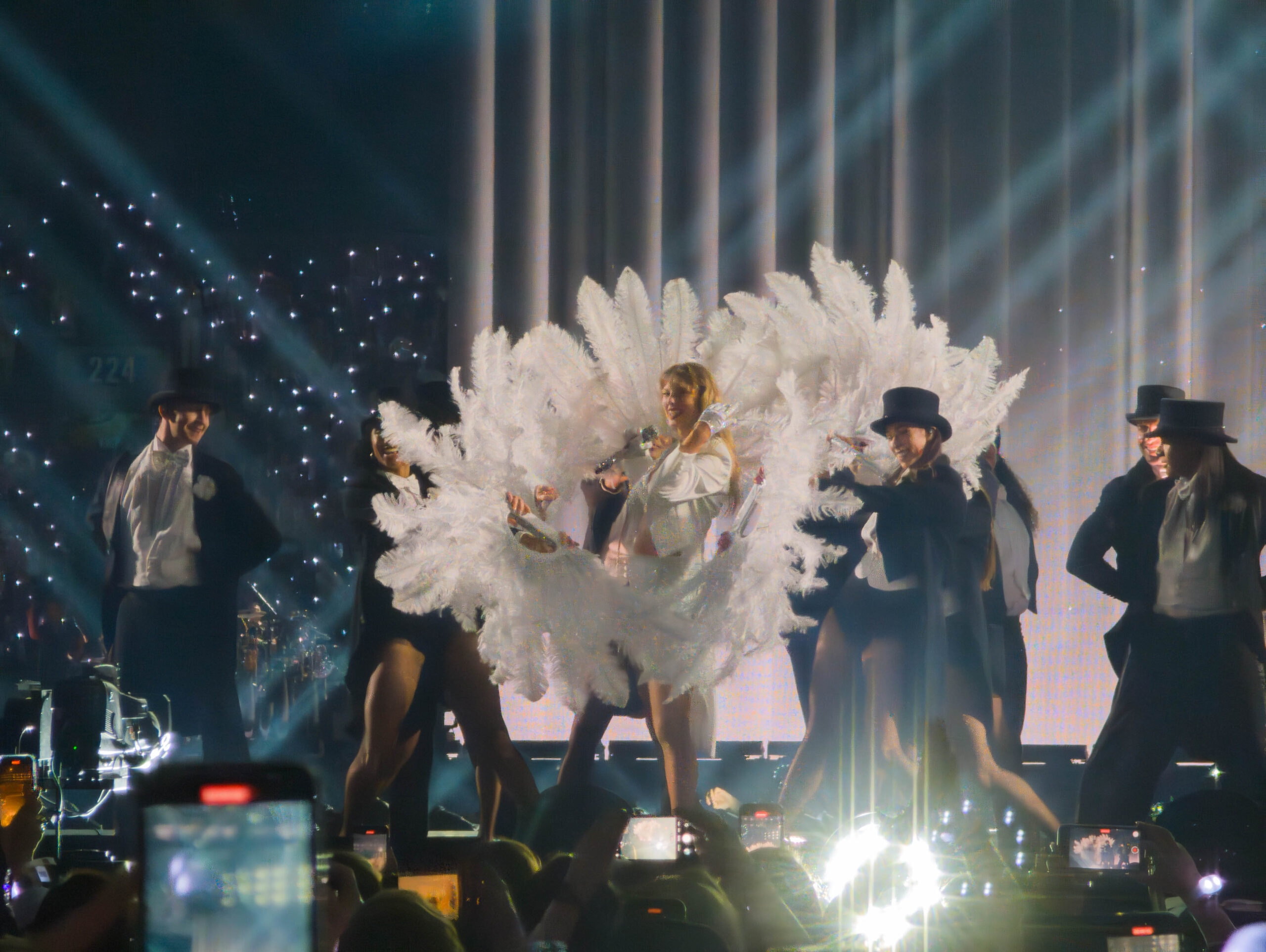 Taylor Swift stands on stage at BC Place surrounded in white feathers and people in top hats.